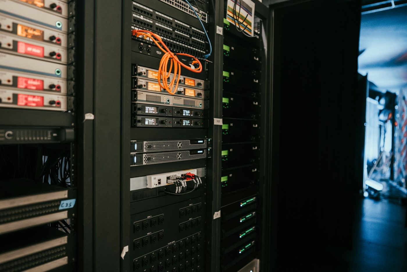 Armarios de equipos electrónicos en una sala a oscuras, con aparatos de pantallas encendidas, cables de red naranjas y azules y paneles de conexiones. Foto: Kevin Ache en Unsplash.