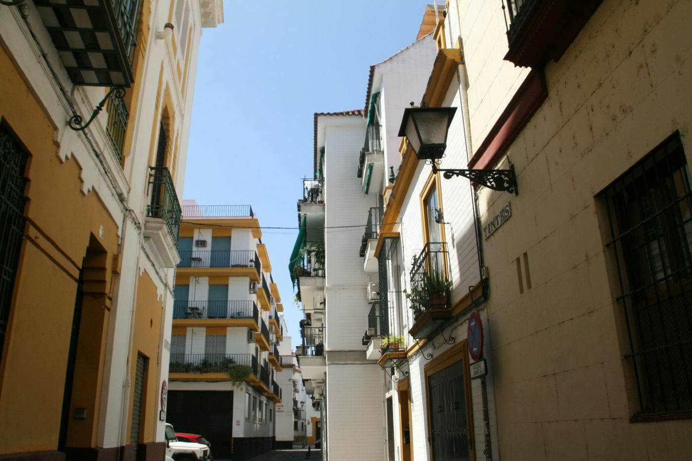 Una calle estrecha del centro de Sevilla, con casas blancas y amarillas, balcones con macetas y un farol en la pared. Foto: Laura Abellán García en Unsplash.