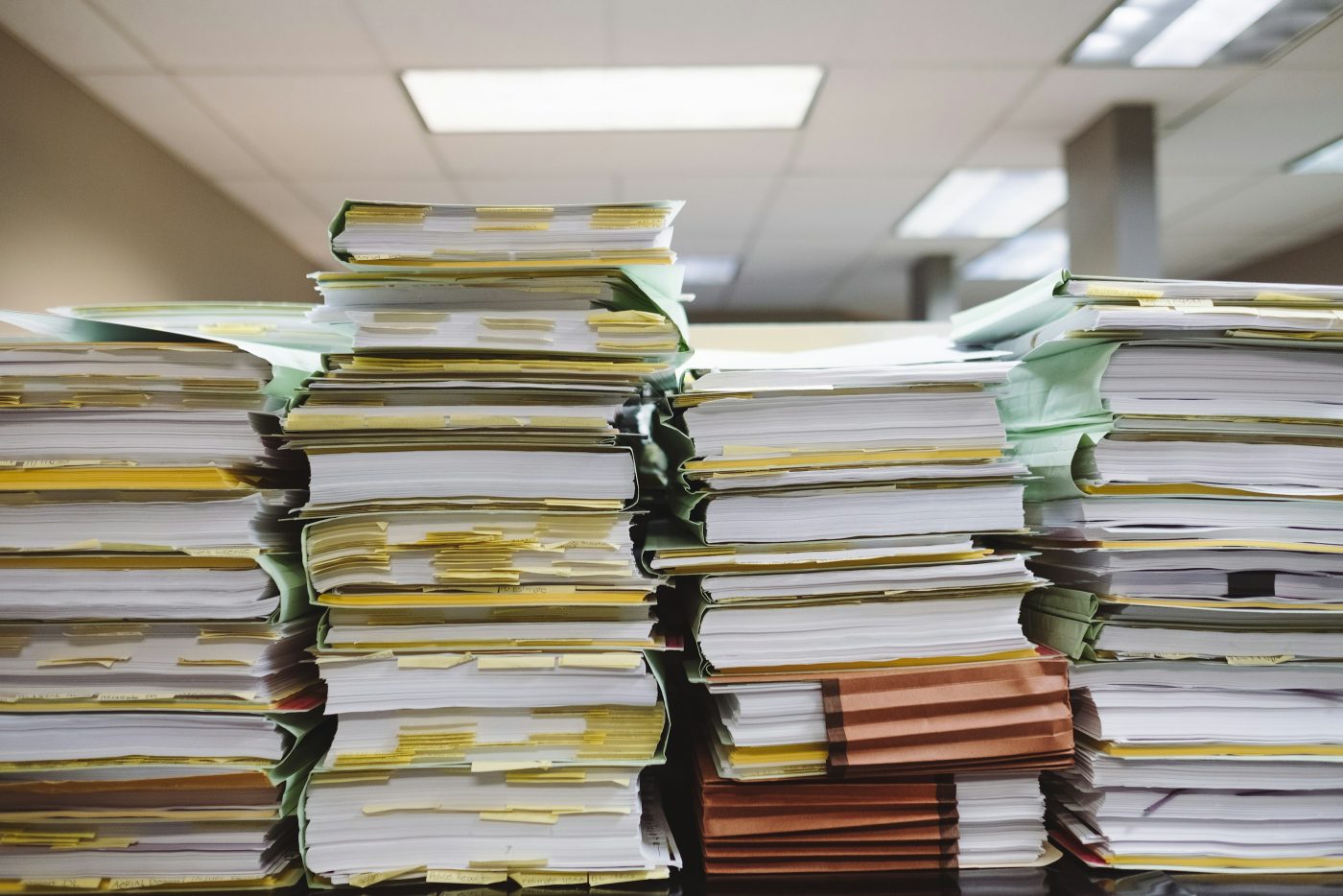 Pilas de carpetas llenas de papeles y notas adhesivas amarillas, en una oficina. Foto: Wesley Tingey en Unsplash.