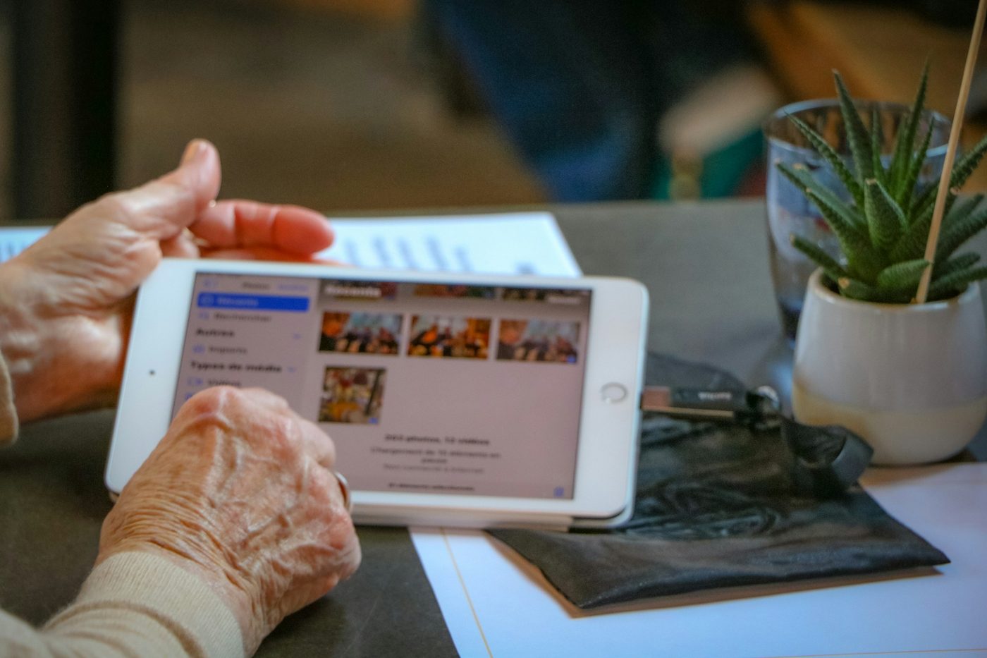 Unas manos de persona mayor recorren la galería de fotos de una tableta, sobre una mesa con papeles y una planta. Foto: Layla Ait Laaraj en Unsplash.