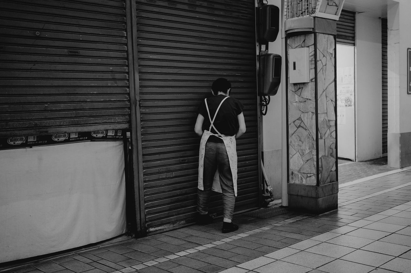 Una persona con delantal, junto a la persiana a medio bajar de una tienda. Foto: Johnny Ho en Unsplash.
